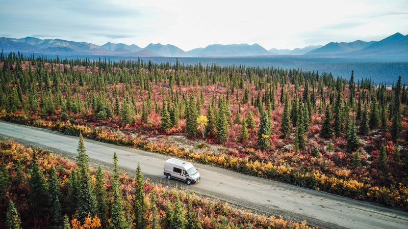 An RV rental travels through a vibrant fall forest, rich with green, red, and orange foliage, against distant mountains. Ideal road trip. — photo 8