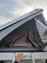 Looking up at an open James Baroud rooftop tent on a vehicle, with a hand gripping the ladder. Ideal for an rv rental.