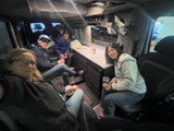 Four people relax in their RV rental, showcasing the compact and functional interior with a kitchen counter and overhead storage.