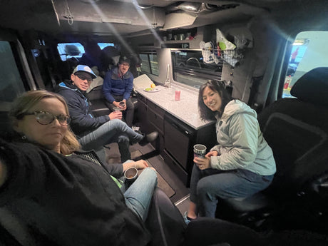Four people relax in their RV rental, showcasing the compact and functional interior with a kitchen counter and overhead storage.