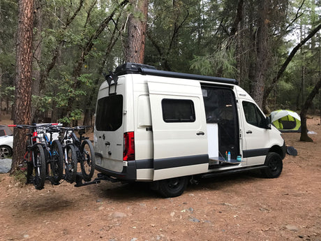 White rv rental camper van with mountain bikes parked at a forest campsite. The side door is open, revealing the interior.