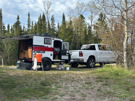 Scenic RV rental: White pickup & teardrop camper with open kitchen, parked in a forest setting for outdoor adventure.
