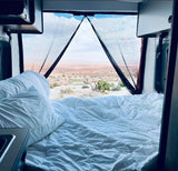 Inside a comfy rv rental, a bed with white linens faces a stunning desert landscape through a mesh window screen, perfect for van life.