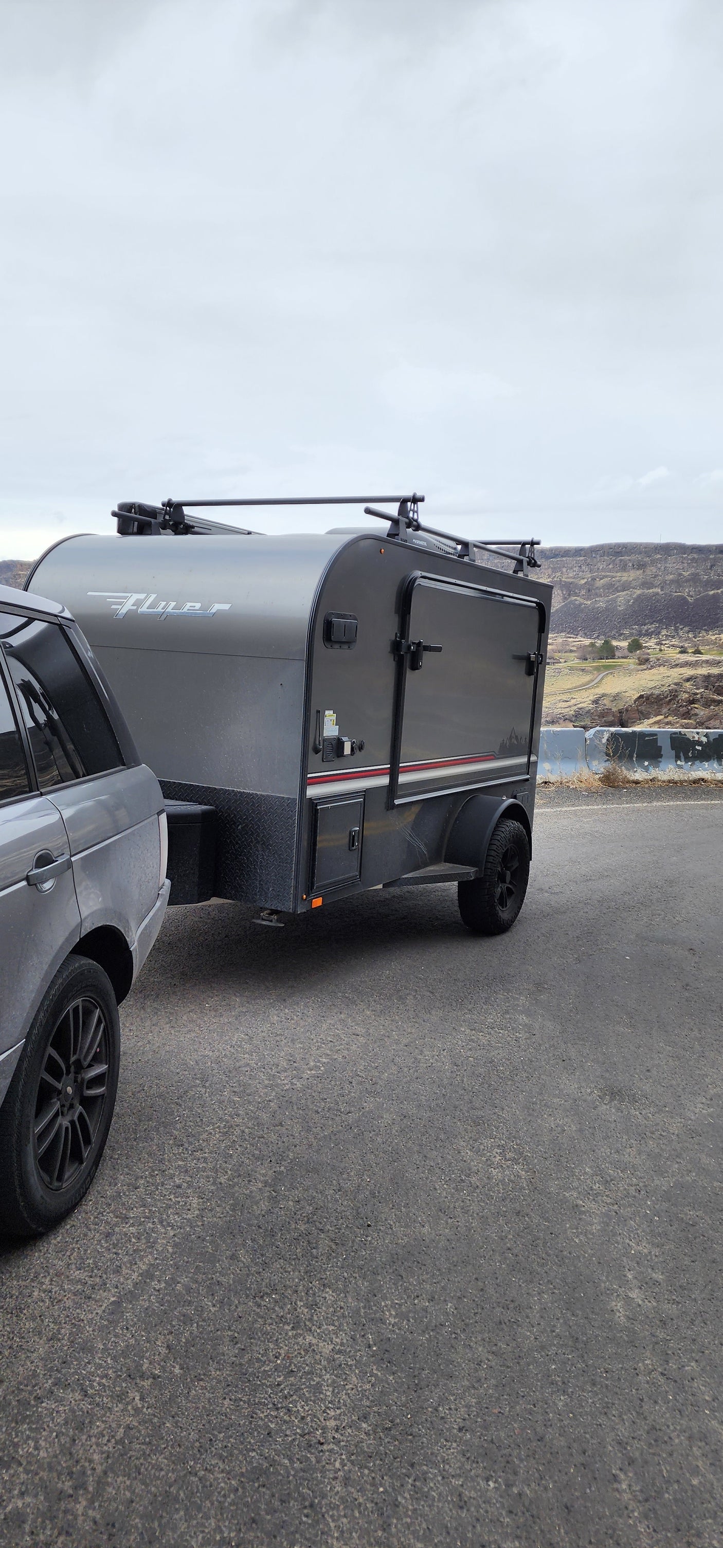 RV rental ready for adventure: Gray SUV towing a "Flyer" teardrop camper with roof racks against a scenic hilly backdrop.
