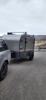 RV rental ready for adventure: Gray SUV towing a "Flyer" teardrop camper with roof racks against a scenic hilly backdrop.