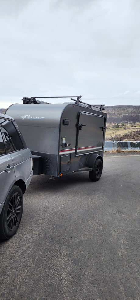 RV rental ready for adventure: Gray SUV towing a "Flyer" teardrop camper with roof racks against a scenic hilly backdrop.