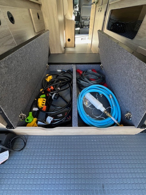 RV rental's rear storage area, neatly organized with cables, adapters, and a water hose in underfloor compartments.
