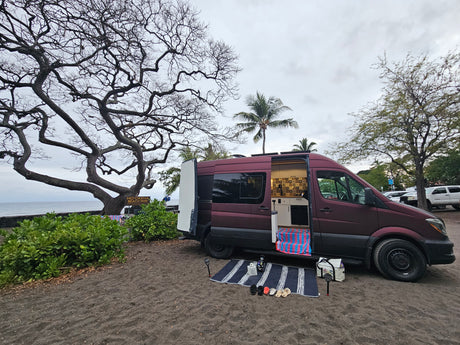 Maroon RV rental van at Ho'okena Beach Park, with its open side revealing a custom kitchen setup & beach mat.
