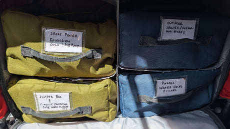 Organized RV rental accessories: labeled bags hold shore power, jumper box, outdoor shower, and potable water hoses & filter.