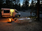 Cozy RV rental camping setup at dusk: camper van, person by campfire, picnic table, and warm string lights.