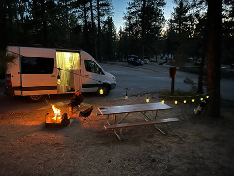 Cozy RV rental camping setup at dusk: camper van, person by campfire, picnic table, and warm string lights.