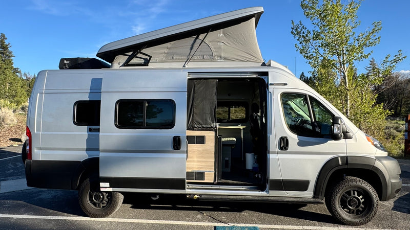  and a coastal tunnel on a sunny day.|Experience nature with this RV rental! A silver camper van is parked by a serene river with mountains and green trees under a bright sky.|Silver pop-top camper van rv rental parked outdoors — photo 3