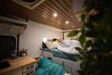 Inside a cozy RV rental: two people sleeping in a modern camper van with wood ceiling & warm lights. Ideal for adventures.
