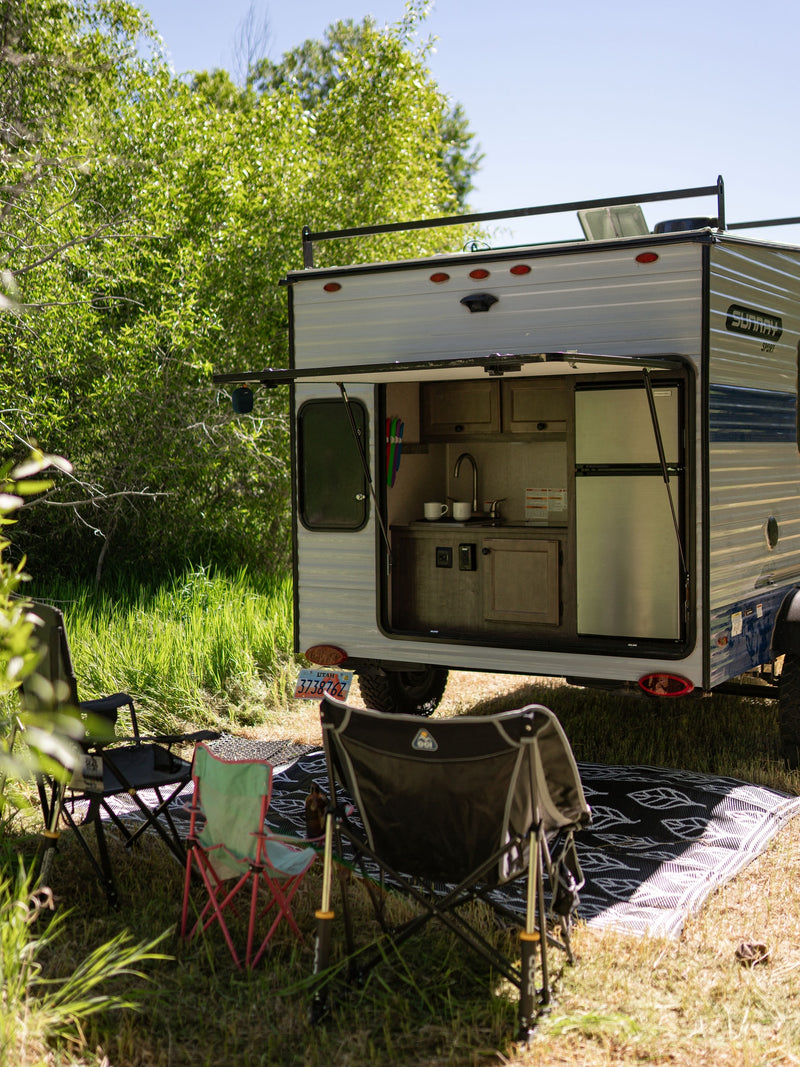 RV rental: Small camper with an open outdoor kitchen, chairs, and mat, set up at a sunny campsite amidst green foliage. — photo 2