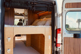 View from the back of an RV rental: a raised bed, modern kitchen, and front seats inside. Open doors reveal a palm tree outside.