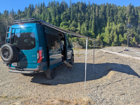 Adventure-ready RV rental: a teal camper van with an awning deployed on a sunny gravel shore by a forest.