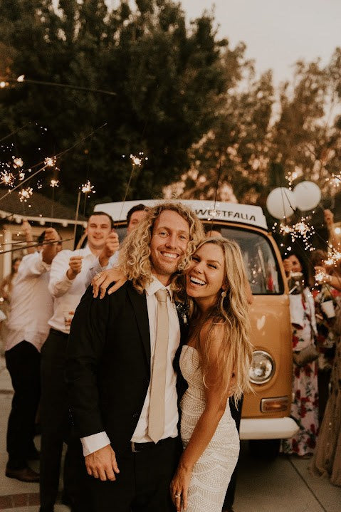 A happy couple poses in front of a vintage orange Westfalia RV rental, surrounded by guests holding sparklers. — photo 18