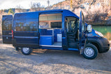 Dark blue RV rental camper van, doors open, showing custom interior: bed, kitchen, fridge, & driver's area. Parked by river.
