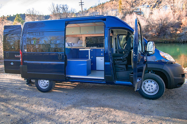 Dark blue RV rental camper van, doors open, showing custom interior: bed, kitchen, fridge, & driver's area. Parked by river.