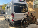 White Ford Transit camper van, equipped with a bike rack holding two mountain bikes, parked on a driveway. Ideal rv rental.