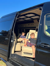 Two boys relax inside a premium RV rental van, showcasing its plush seating and custom interior via an open side door.