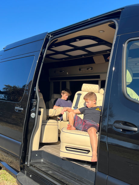 Two boys relax inside a premium RV rental van, showcasing its plush seating and custom interior via an open side door.