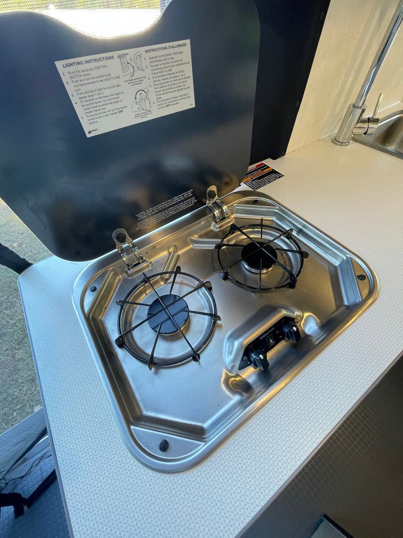 Compact RV rental kitchen featuring a two-burner stainless steel cooktop, open lid with lighting instructions, and a sink. — photo 6