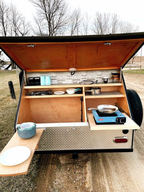 Compact outdoor kitchen in an RV rental teardrop trailer, set up for camping with stove, dishes, and pull-out counter.