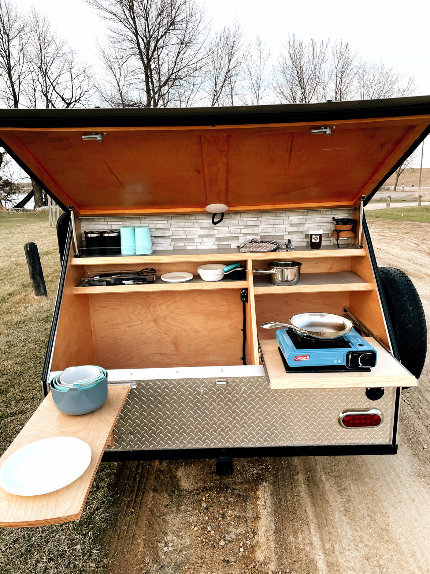 Compact outdoor kitchen in an RV rental teardrop trailer, set up for camping with stove, dishes, and pull-out counter.