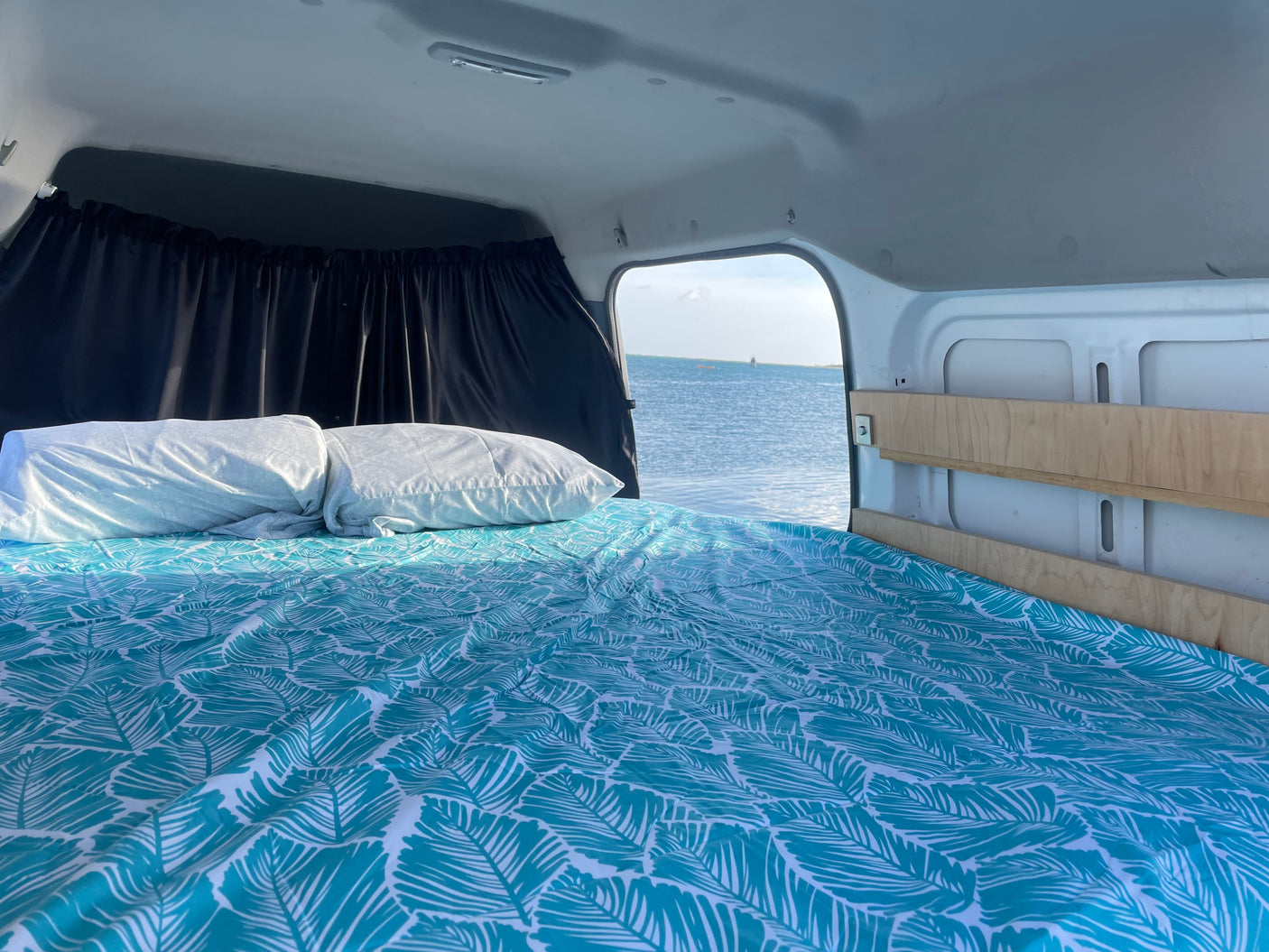 Inside a bright RV rental: a bed with a teal leaf-patterned duvet and white pillows overlooks a calm ocean. Ideal campervan adventure.