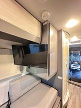 RV rental's cozy interior, showing a wall-mounted TV, comfy sofa, and overhead storage. Smoke detector visible on ceiling.