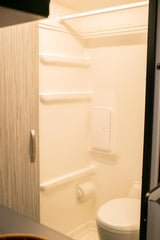 Interior of an RV rental wet bath, showing a white toilet, built-in shelves, shower rod, and toilet paper holder.