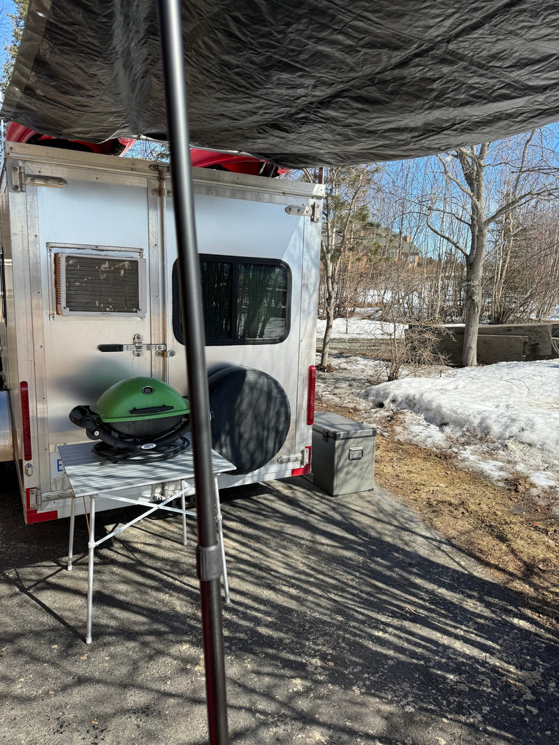 RV rental setup: A silver trailer with a green grill, cooler, and tarp awning. Parked in a snowy, wooded area for outdoor adventure. — photo 6