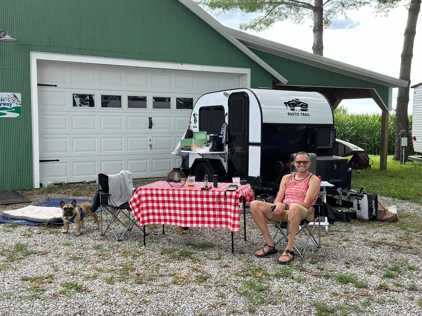 Man & dog relax by a Koala teardrop **RV rental** with picnic table setup at a rustic campsite.
