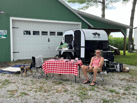 Man & dog relax by a Koala teardrop **RV rental** with picnic table setup at a rustic campsite.