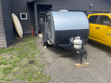 Grey teardrop camper RV rental on gravel, with propane tank, parked beside a dark building & yellow pickup. Surfboard visible.