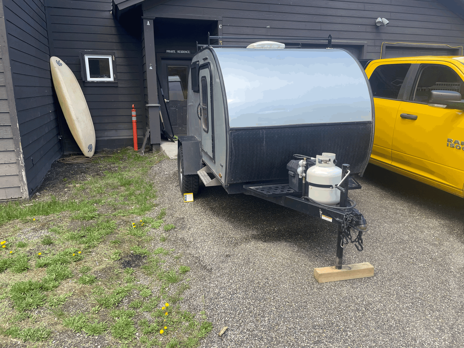 Grey teardrop camper RV rental on gravel, with propane tank, parked beside a dark building & yellow pickup. Surfboard visible.