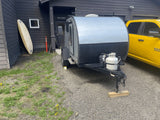 Grey teardrop camper RV rental on gravel, with propane tank, parked beside a dark building & yellow pickup. Surfboard visible.
