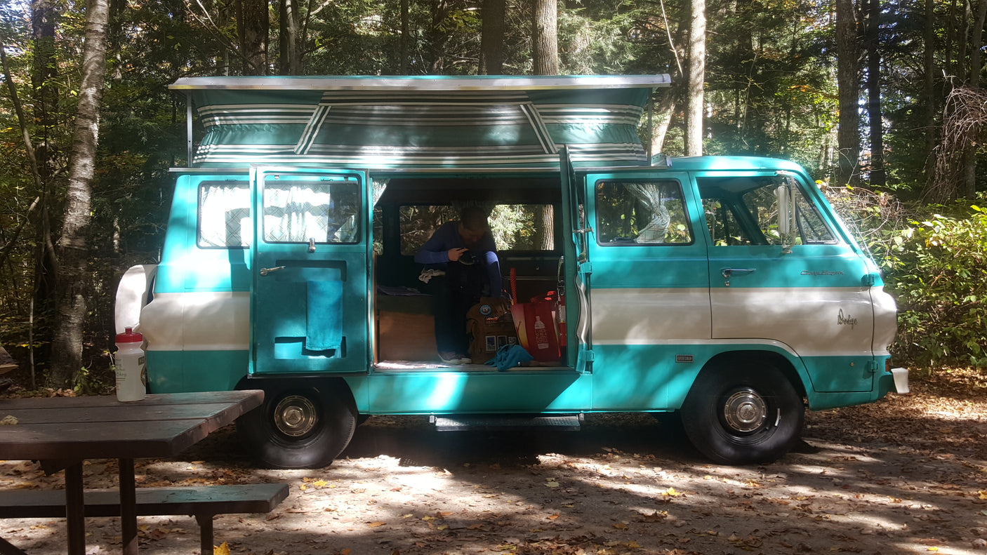 Vintage teal & white pop-top camper van at a wooded campsite with a person inside. Ideal for an RV rental adventure.