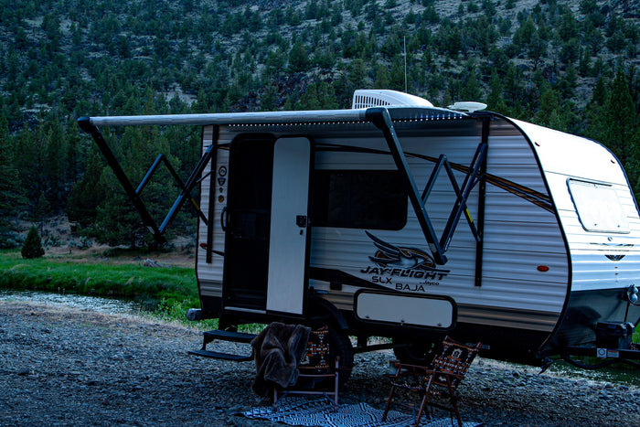  and two happy dogs at a beautiful outdoor campsite.|A truck towing a light travel trailer (Jayco Jay Flight SLX Baja) in front of an American flag mural. Perfect for your next rv rental trip!|An RV rental Jayco trailer and black truck parked roadside with an extended awning by a river & wooded mountain.|Riverside RV rental campsite at dusk