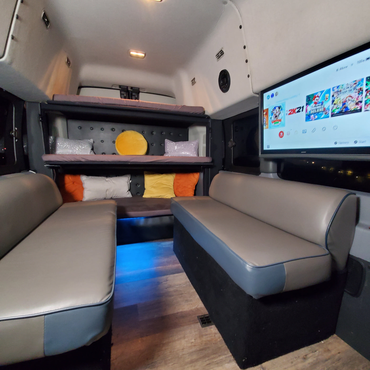 Inside a unique RV rental: two lounge sofas, bunk beds, a large TV for gaming, and cool blue ambient lighting.