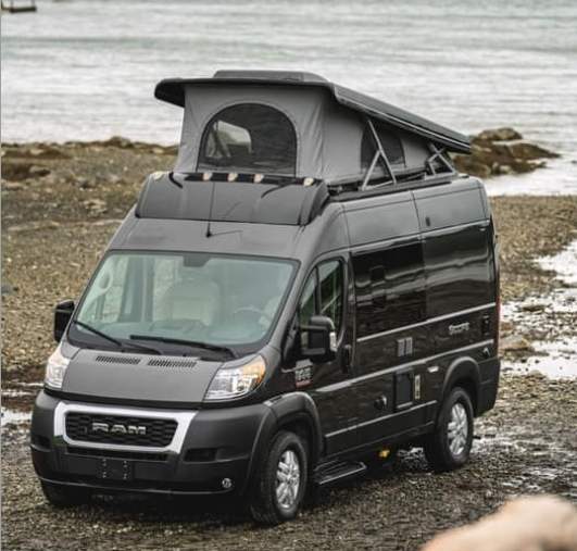 A dark grey Ram ProMaster camper van with an extended pop-top roof, ideal for RV rental, parked on a rocky beach.