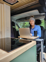 Smiling woman remote working in a cozy RV rental with a laptop, cooktop, & forest views.