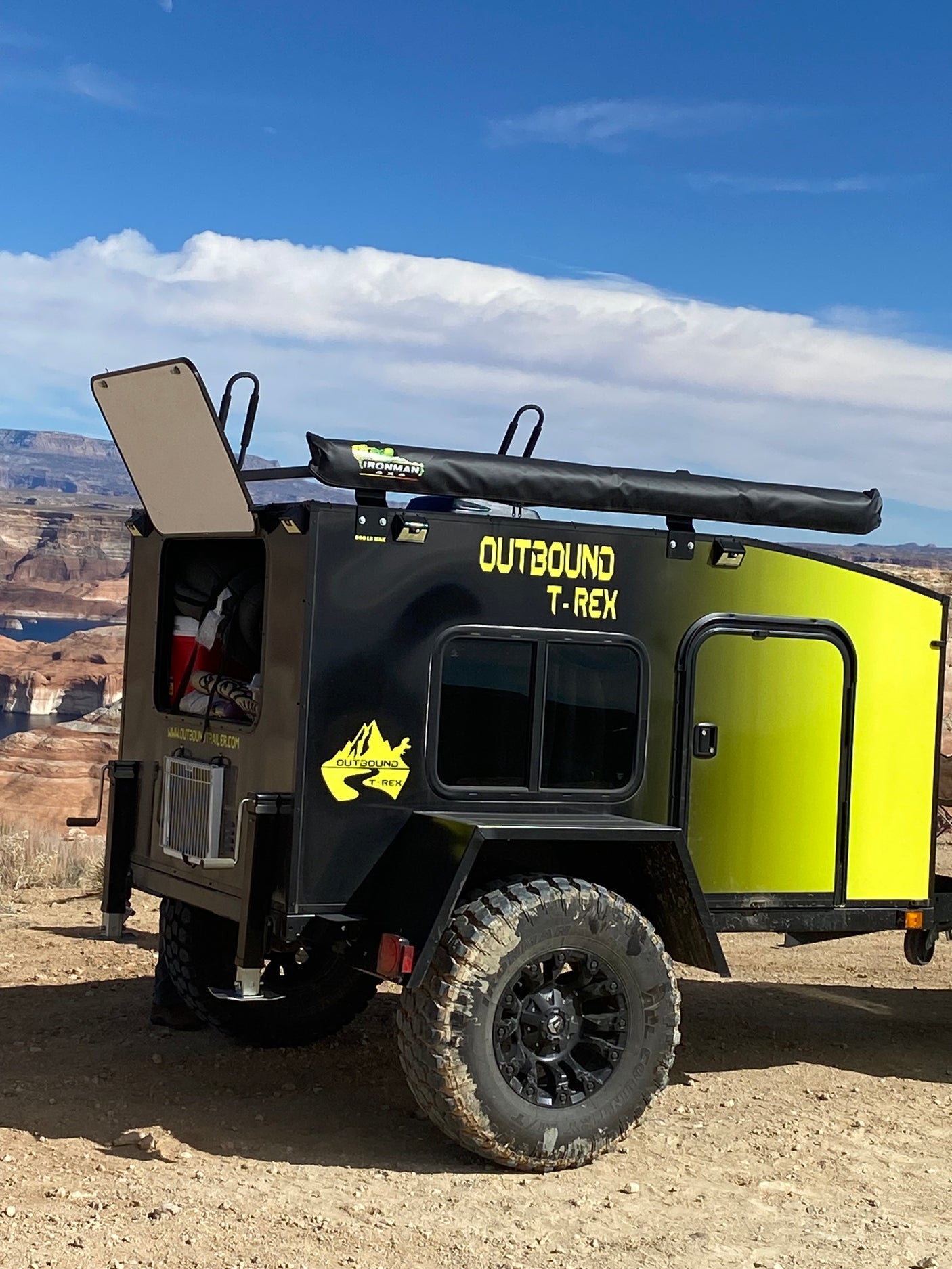 A rugged "Outbound T-Rex" off-road trailer, ideal for an RV rental adventure, parked by a scenic lake with desert mountains.