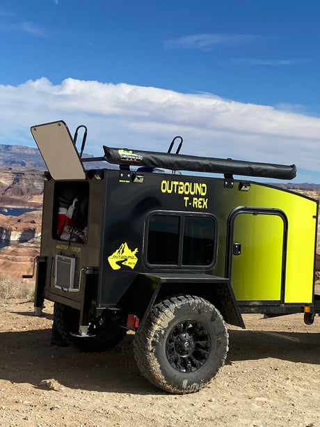 A rugged "Outbound T-Rex" off-road trailer, ideal for an RV rental adventure, parked by a scenic lake with desert mountains.