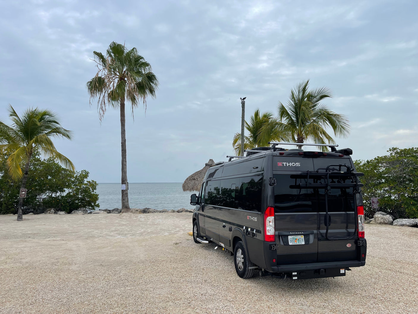 A dark gray Ethos camper van is parked on gravel near tropical water under cloudy skies, ready for an RV rental adventure.