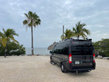 A dark gray Ethos camper van is parked on gravel near tropical water under cloudy skies, ready for an RV rental adventure.