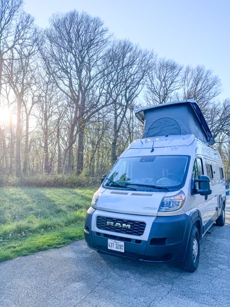 An RV rental Ram camper van with its pop-top roof extended is parked on a road, bathed in sunrise light through trees.