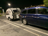 Night scene: Blue minivan towing a teardrop travel trailer in a parking lot, ready for an RV rental adventure.
