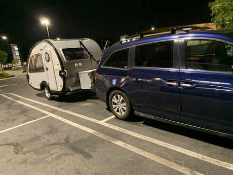 Night scene: Blue minivan towing a teardrop travel trailer in a parking lot, ready for an RV rental adventure.